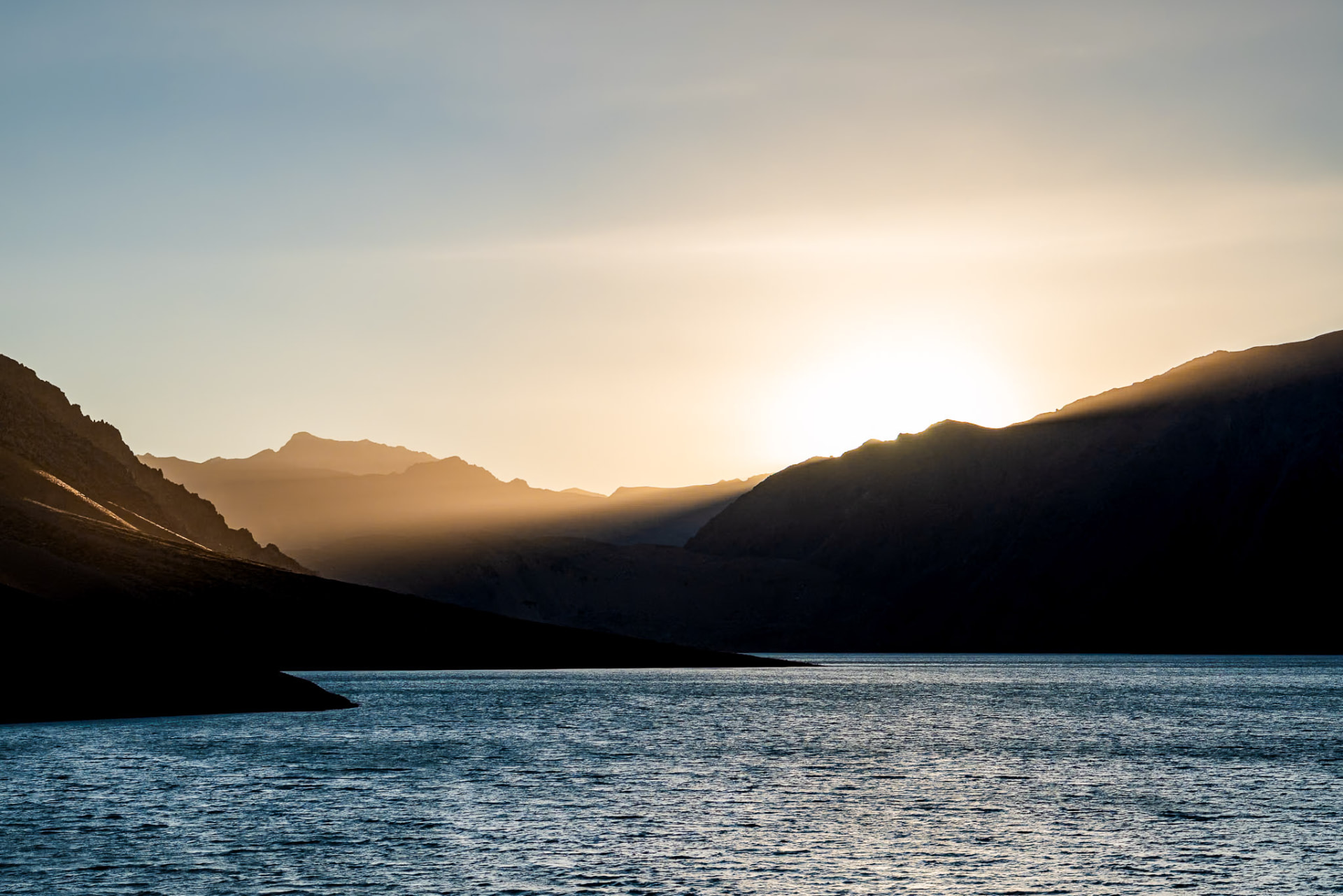 Masa de agua del Embalse El Yeso rodeada por perfiles de cerros con el sol escondiéndose detrás